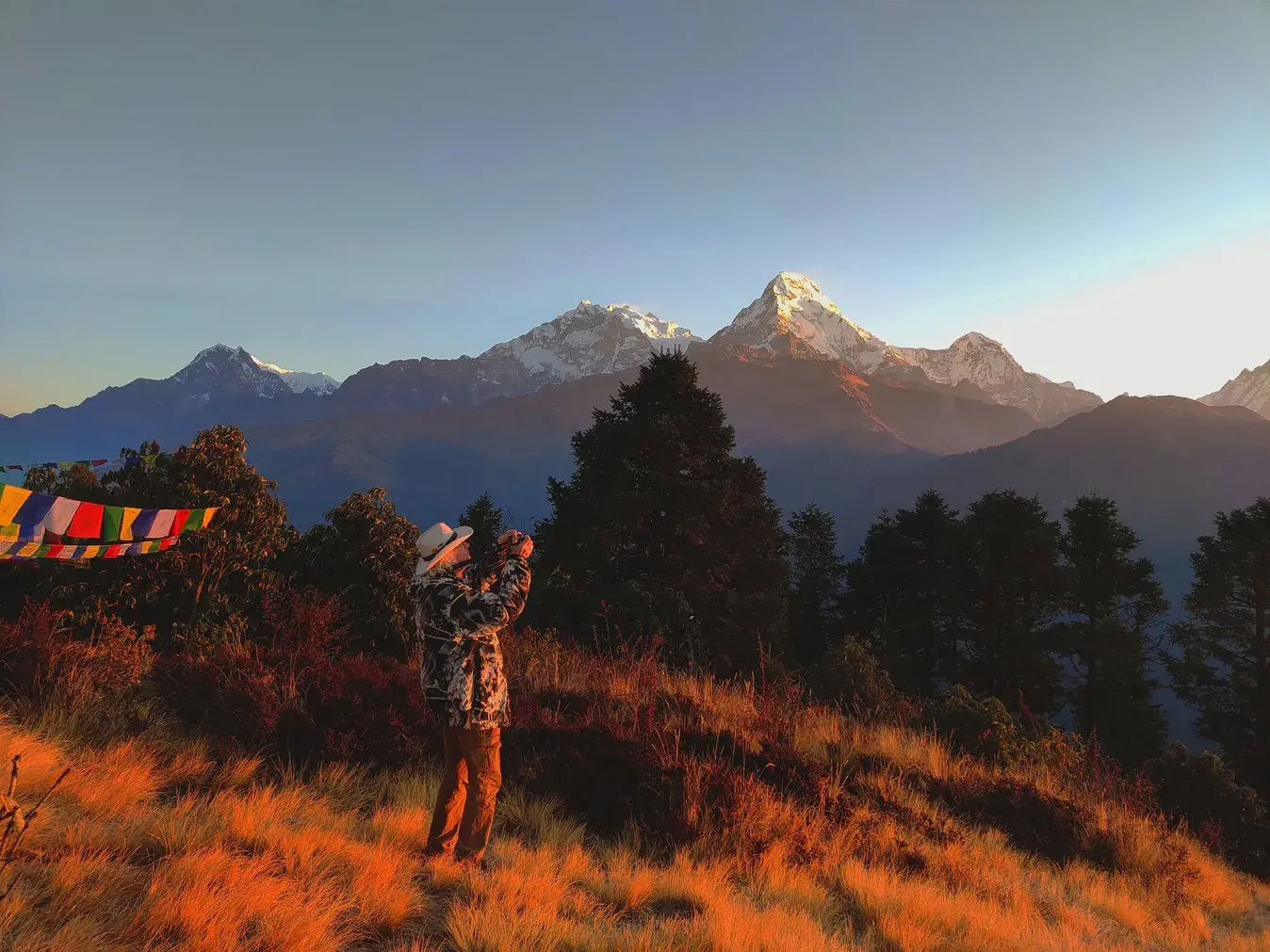 Poon Hill Trek- 3 Days A person wearing a hat stands on a golden grassy hill taking a picture of snow-covered Himalayan mountain peaks at sunrise, with colorful prayer flags visible on the left.