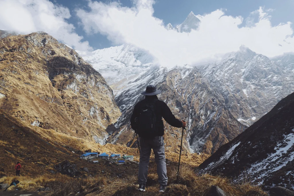 A hiker with a backpack and trekking poles looking out over a mountain valley and snowy peaks.