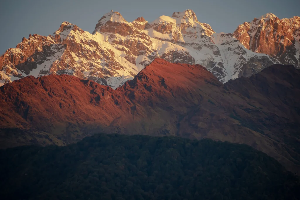 Snow-capped mountain peaks glowing in warm golden sunlight above dark rocky ridges during sunrise on the Annapurna Base Camp Trek Elevation