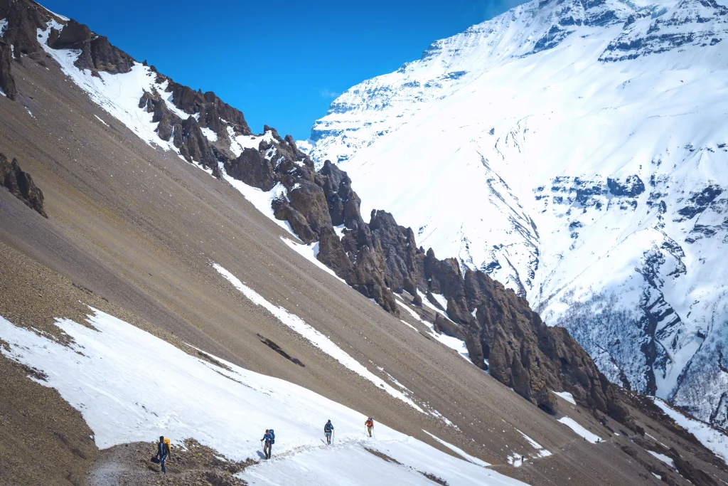 Four hikers with backpacks walking on a snowy mountain trail in the Himalayas.