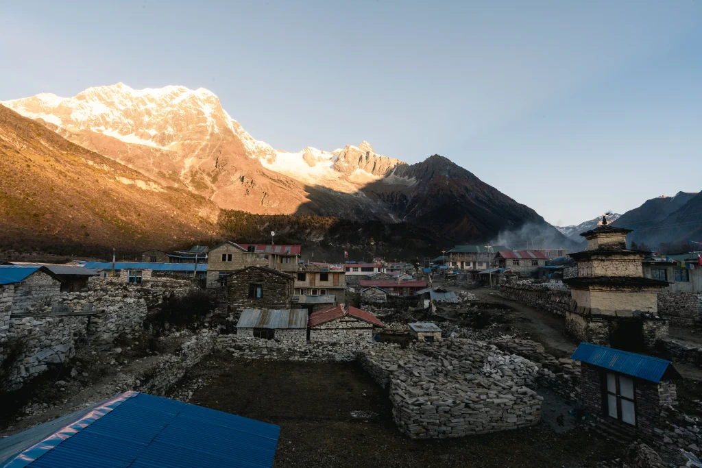 A mountain village with stone houses and a stupa, backed by snow-covered peaks catching the morning light.