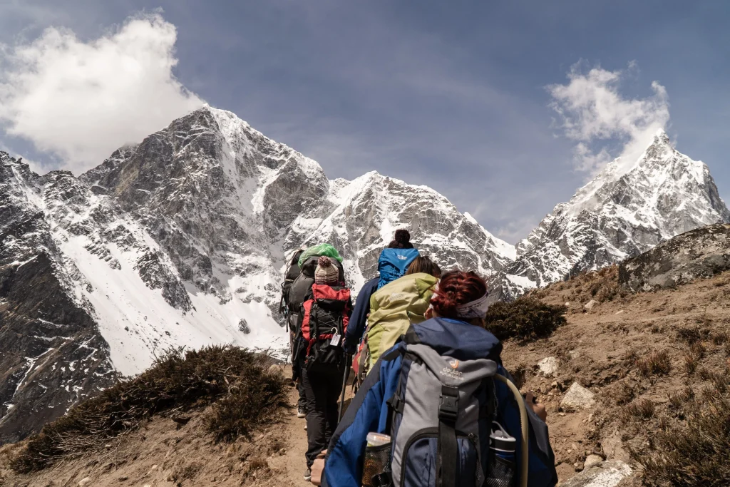 A group of trekkers carrying large backpacks walking on a mountain trail with snow-covered peaks in the background.