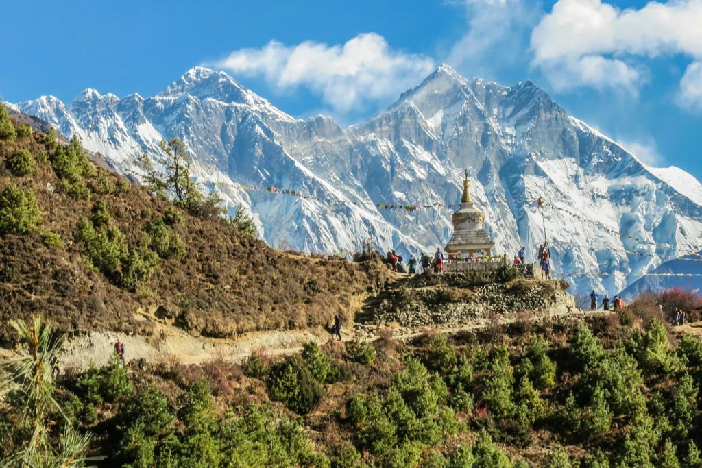 A mountain hillside with green trees and a stupa, backed by towering snow-capped peaks.