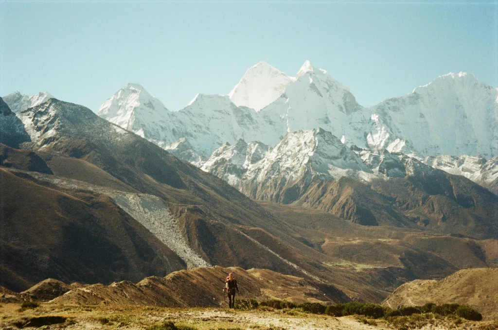 A lone trekker with a backpack and trekking poles walking along a high-altitude ridge toward snowy mountain peaks on the everest base camp trek.