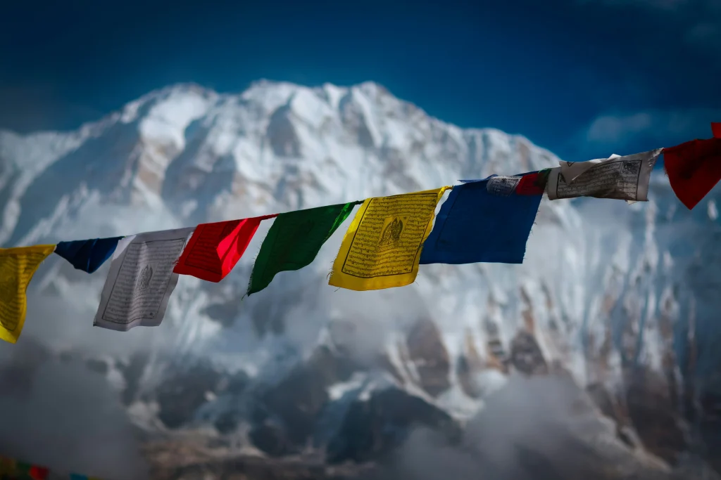A line of colorful Buddhist prayer flags stretched across the foreground with a massive, snow-covered Himalayan peak in the blurred background under a deep blue sky.