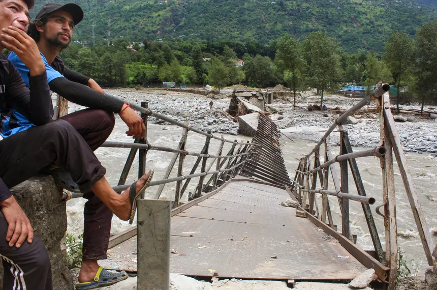 Two men sitting near a collapsed metal pedestrian bridge destroyed by flash floods over a rocky mountain river, showing trail damage along a trekking region.