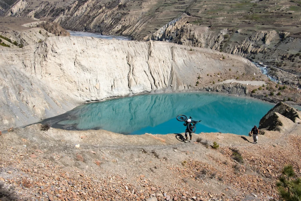 A trekker on the Annapurna Circuit carrying a mountain bike up a steep path next to a vibrant turquoise glacial lake and traditional stone monastery