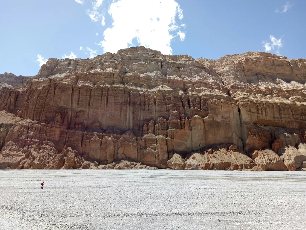A lone figure walking along a wide, dry gravel riverbed in front of massive, eroded rock cliffs near Kagbeni on the Annapurna Circuit.