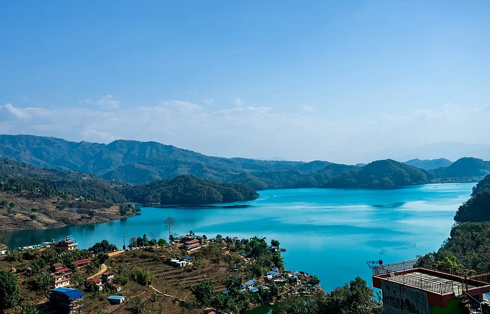 Vibrant turquoise waters of Begnas Lake in Pokhara surrounded by green hills and village settlements under a clear blue sky.