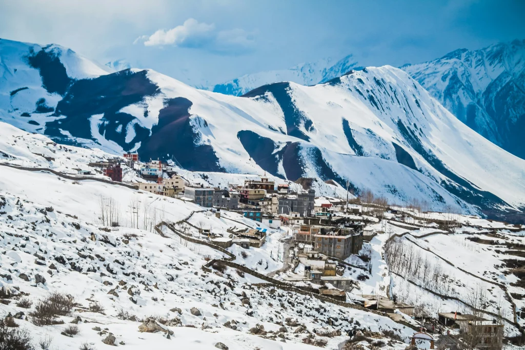 A wide view of the snow-blanketed village of Muktinath nestled on a hillside along the Annapurna Circuit with towering snow-capped Himalayan mountains behind it.