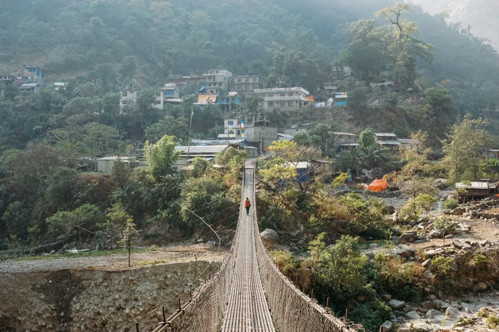 A lone trekker walking across a long steel suspension bridge toward a hillside village along the Manaslu Circuit.