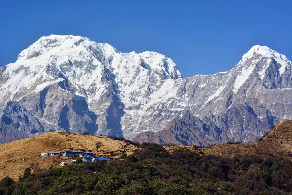 A scenic mountain landscape showing a small cluster of blue-roofed teahouses on a golden hillside nestled below massive, snow-covered Himalayan peaks against a vibrant clear blue sky, illustrating a traditional trekking stop.