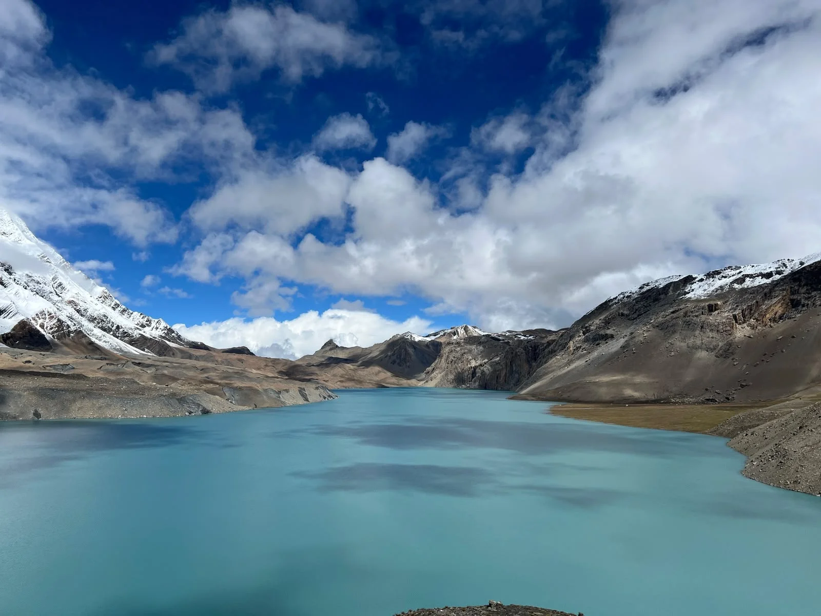 Turquoise-blue Tilicho Lake surrounded by rugged, snow-covered Himalayan mountains beneath a bright blue sky with white clouds.