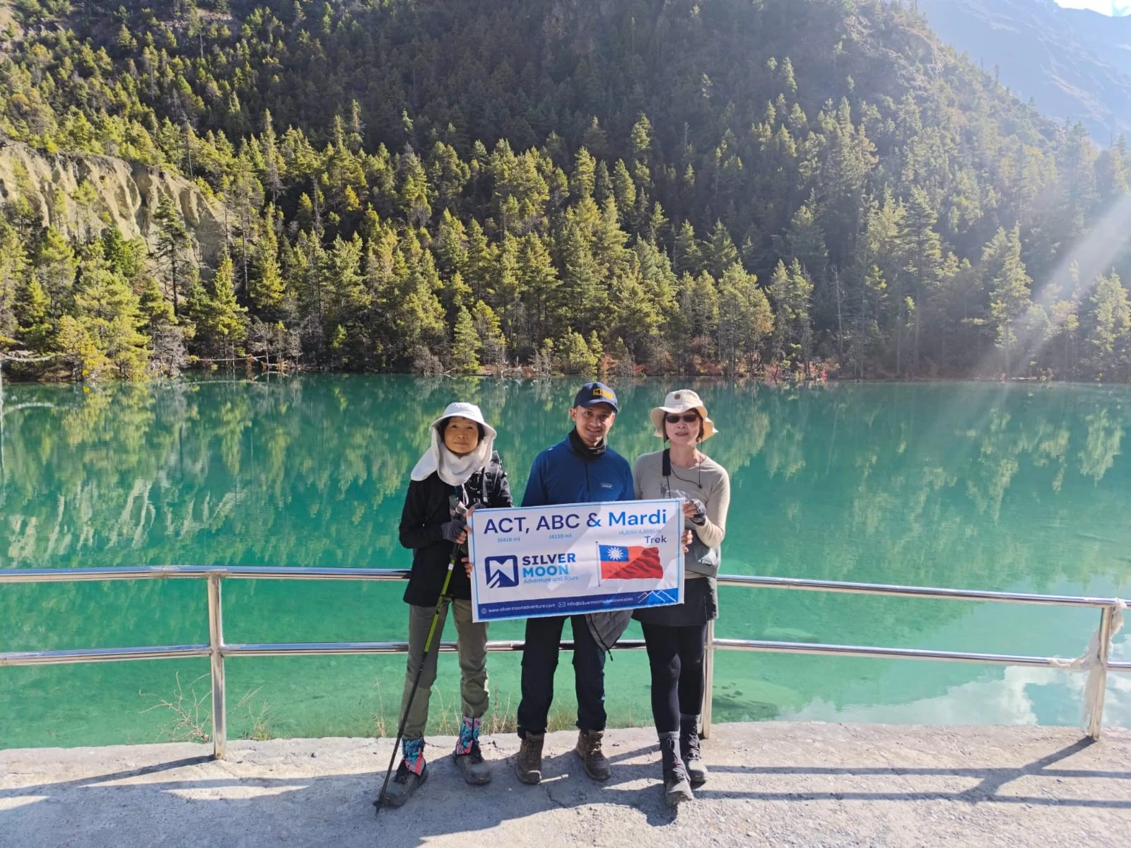 Annapurna Circuit Trek with Nar Phu Valley -27 Days Trekkers holding a Silver Moon Adventure banner beside a turquoise alpine lake surrounded by pine forests in the Himalayas.