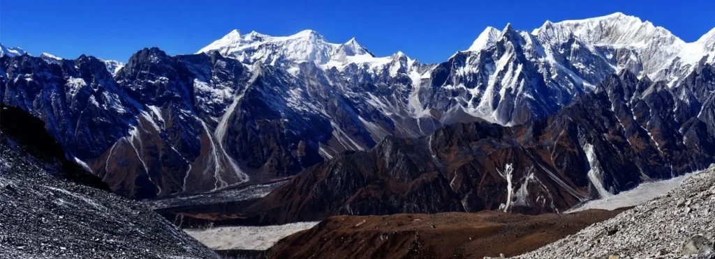 A wide panoramic vista of towering snow-capped Himalayan mountain peaks above deep dark valleys and glaciers under a clear blue sky, highlighting spectacular trekking scenery.