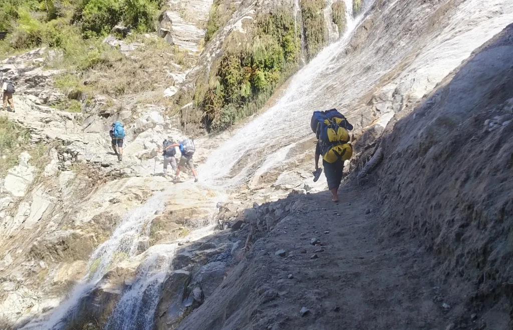 Trekkers carrying heavy backpacks hike up a steep, rocky mountain path crossing past a rushing waterfall along the Manaslu Circuit.