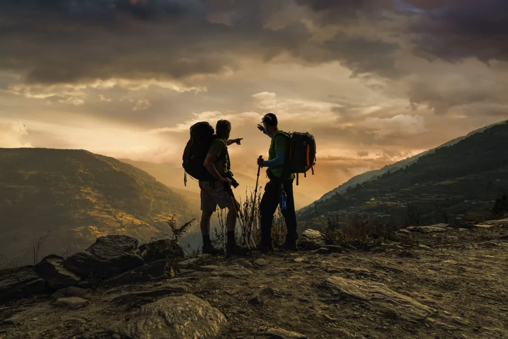 A scenic photograph of two backpackers standing silhouetted on a rocky trail at sunset, pointing across a vast mountain valley.