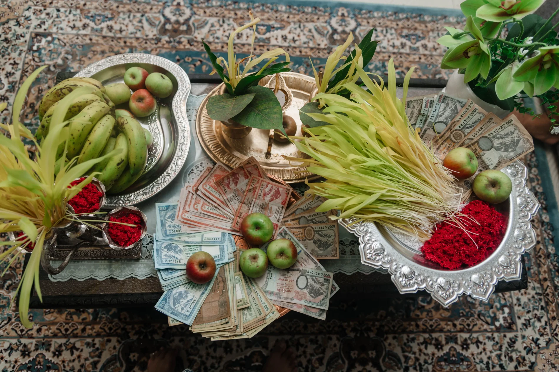 An overhead view of a Dashain festival arrangement with red Tika, yellow Jamara shoots, green bananas, apples, and fanned-out Nepalese rupee banknotes for Dakshina on a patterned table.