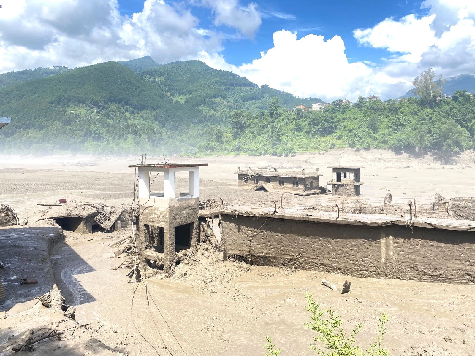 A white concrete watchtower and nearby structures partially buried in a vast expanse of thick mud and flood debris, set against a backdrop of lush green mountains under a cloudy blue sky.