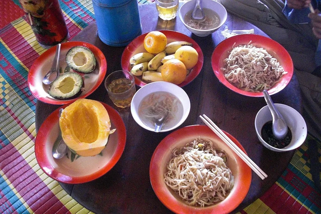 A colorful tabletop spread of tropical fpoood including fresh papaya, avocado, bananas, oranges, noodles, and soup.