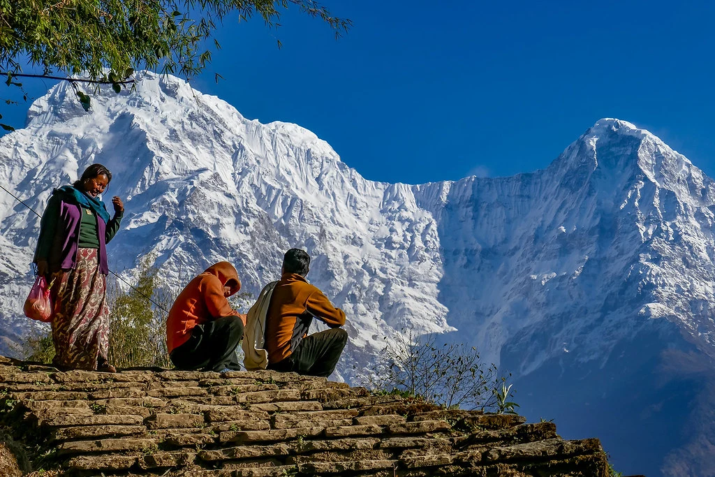 Local residents resting on a steep stone-paved staircase with massive, snow-covered Himalayan peaks rising against a clear blue sky.