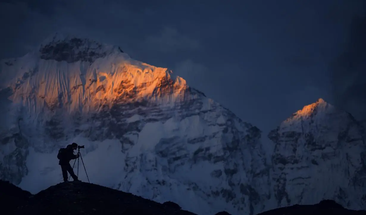 A photograph of a silhouetted photographer with a tripod on a dark ridge in front of snow-covered mountain peaks lit by golden light.