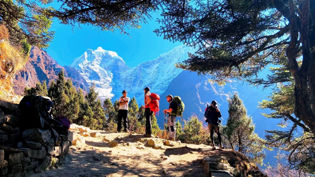A group of trekkers resting along a sunlit mountain trail surrounded by trees and peaks, showcasing the trekking experience in a standard vs luxury ebc trek.