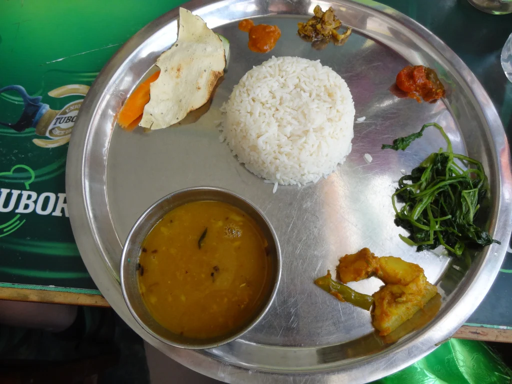 a metal thali plate serving traditional Nepali Dal Bhat, including steamed rice, lentil soup, saag greens, vegetable curry, chutney, and papad