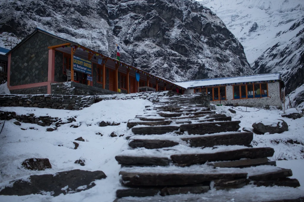 A stone staircase covered in snow leading up to a wooden guest house and restaurant surrounded by steep, snow-dusted rocky mountains.