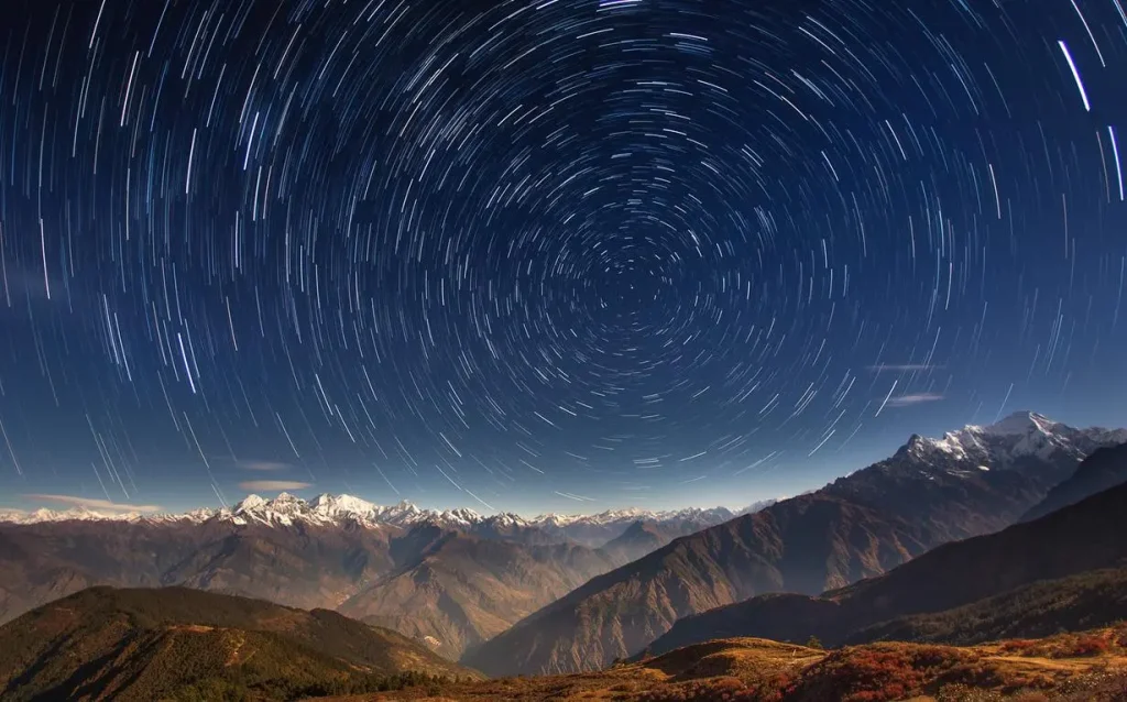 A long-exposure photograph showing circular star trails swirling in the night sky directly above snow-capped mountain peaks and deep valleys