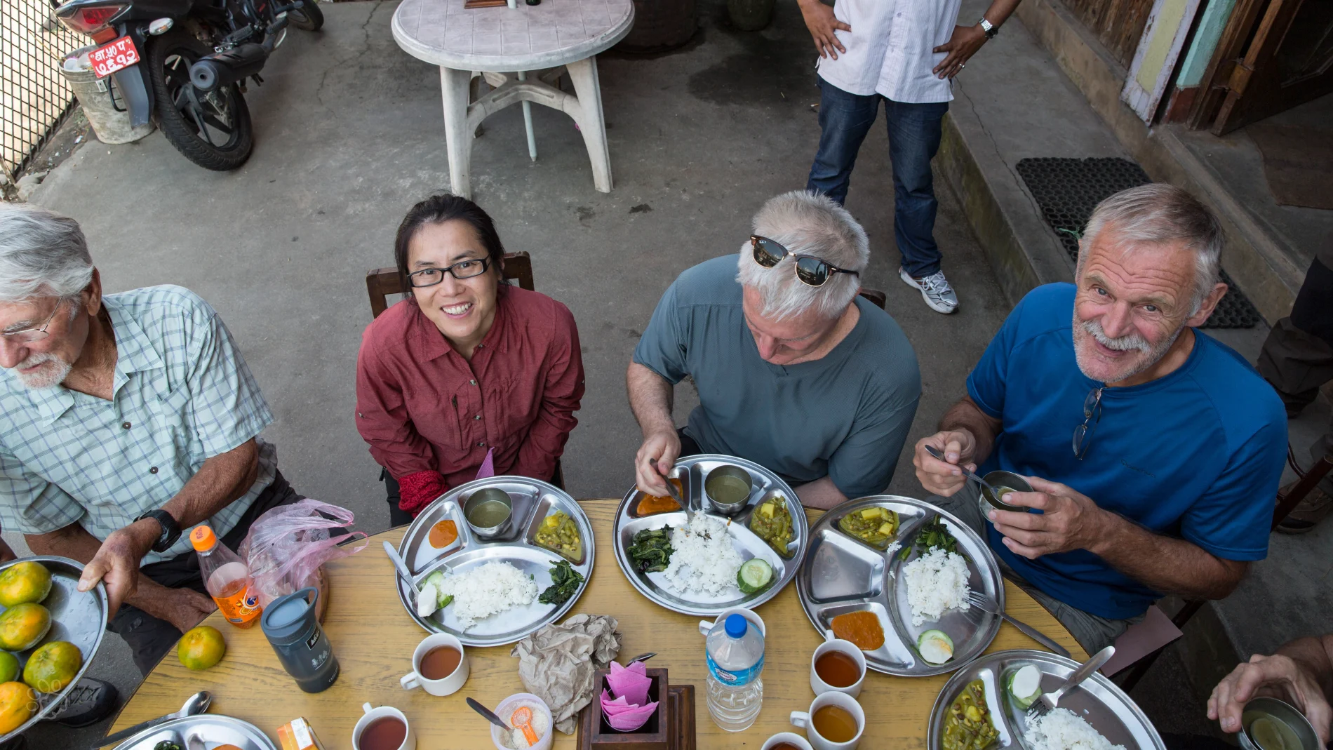 A group of people sitting around an outdoor table sharing a traditional food meal served on stainless steel plates.