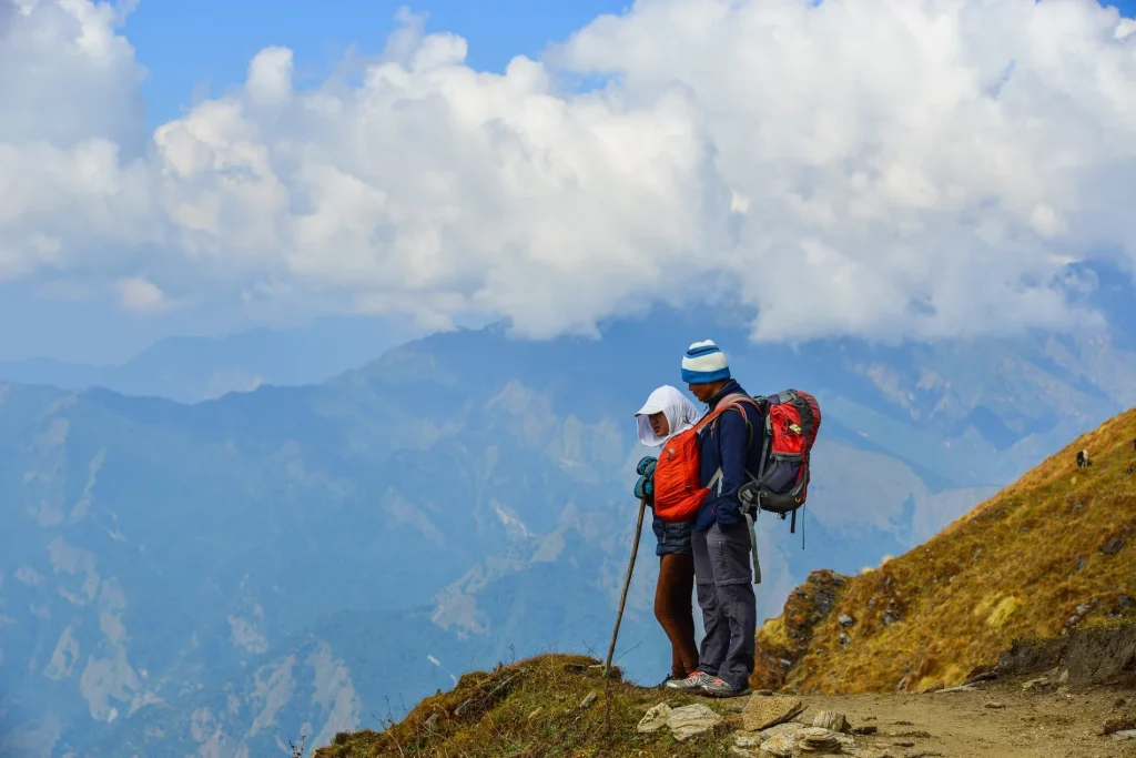 A scenic photograph of two hikers with backpacks standing on a high mountain trail overlooking distant blue ranges under cloudy skies.
