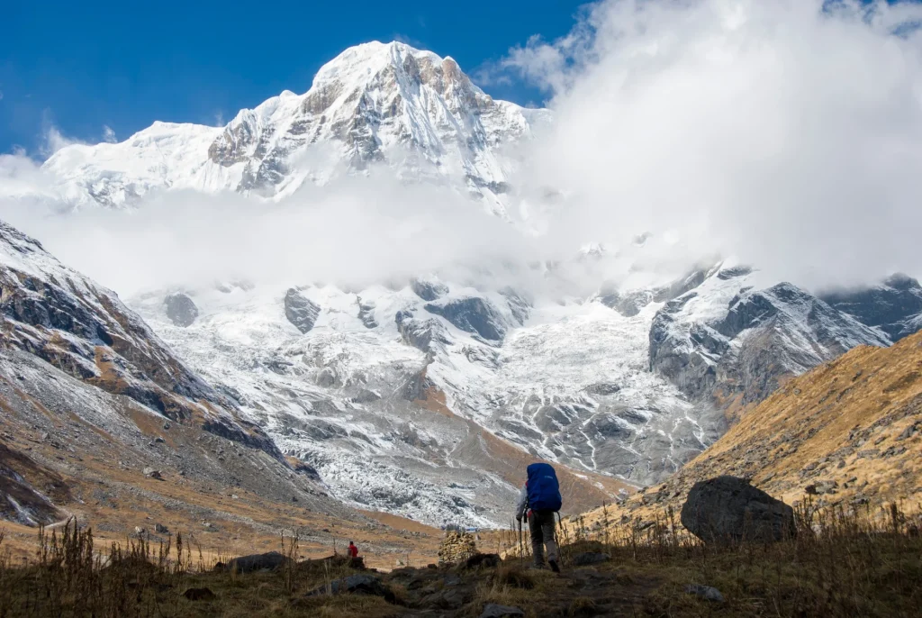 A hiker with a large blue backpack walking along a rugged trail in a high-altitude valley toward a majestic, snow-covered Himalayan mountain peak under a clear blue sky, illustrating a Himalayan trekking adventure.
