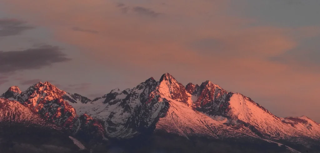 featuring snow-capped mountain peaks bathed in the warm, pinkish-orange light of dawn under a dramatic morning sky