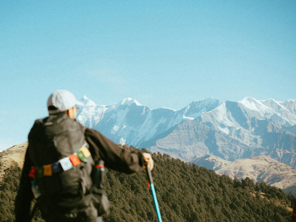 Rear view of a trekker carrying a backpack with prayer flags, using trekking poles while looking out toward snow-capped mountain peaks.