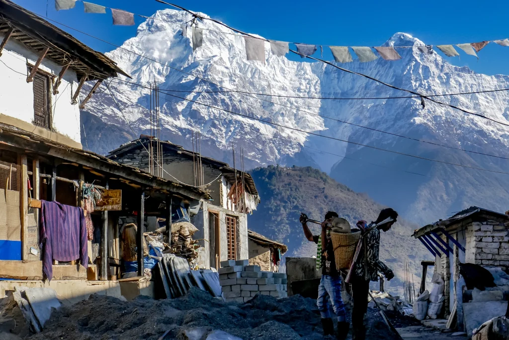 a trekker passing local villagers carrying baskets along a stone path in a Himalayan village set against massive snow-capped peaks.